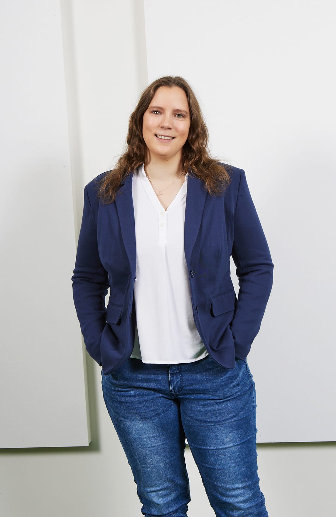 Eine l&auml;chelnde Frau mit langen, lockigen Haaren tr&auml;gt einen blauen Blazer und steht vor einem hellen Hintergrund.