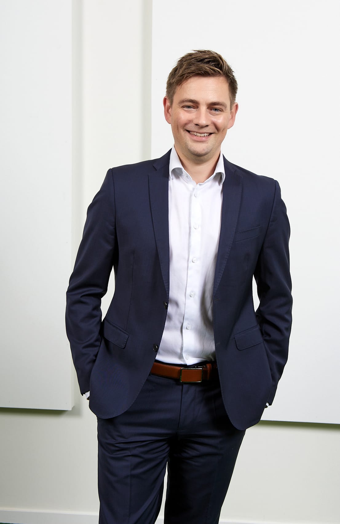 Ein lächelnder Mann in einem dunkelblauen Anzug steht mit verschränkten Händen vor einer weißen Wand.