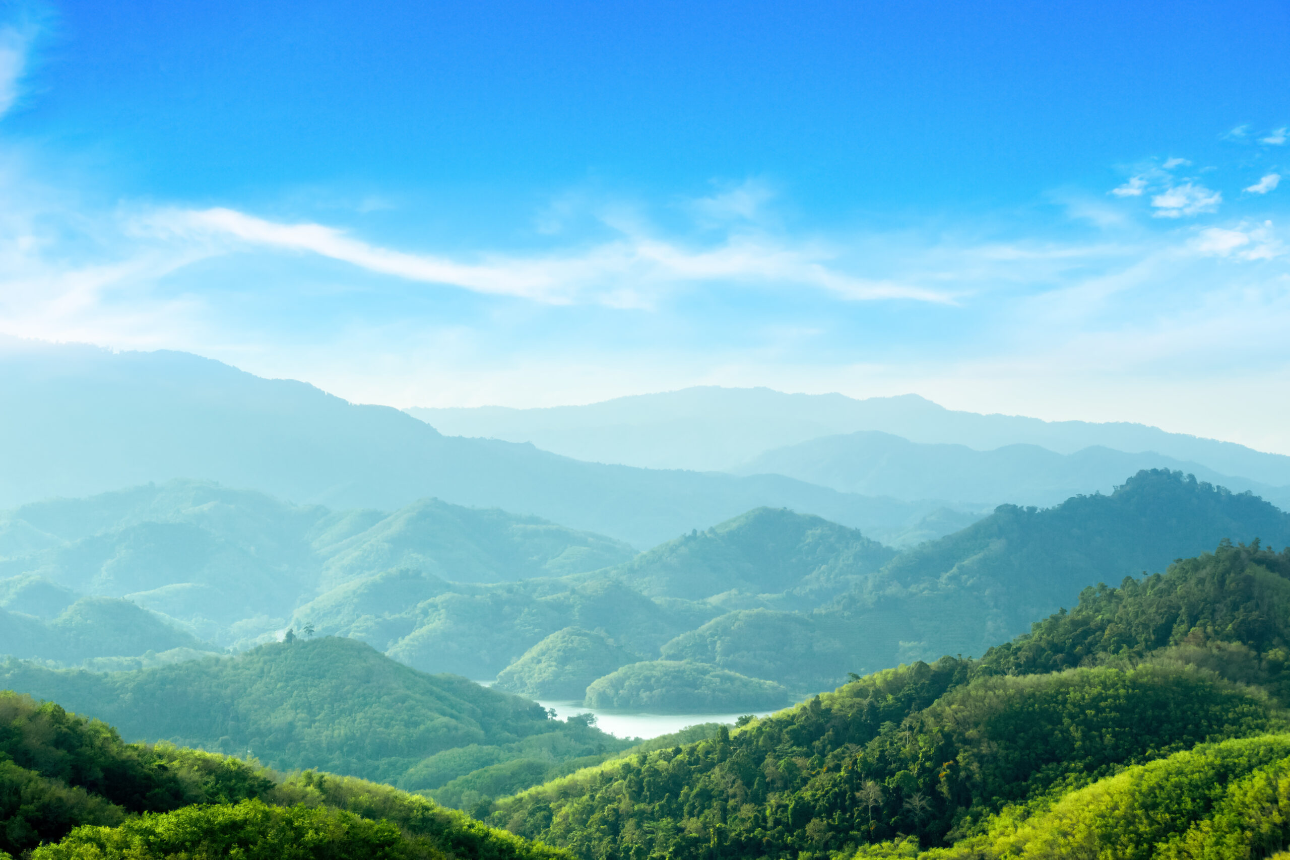 Blick auf gr&uuml;ne H&uuml;gel und Berge unter einem klaren blauen Himmel, mit einem schimmernden See im Hintergrund.