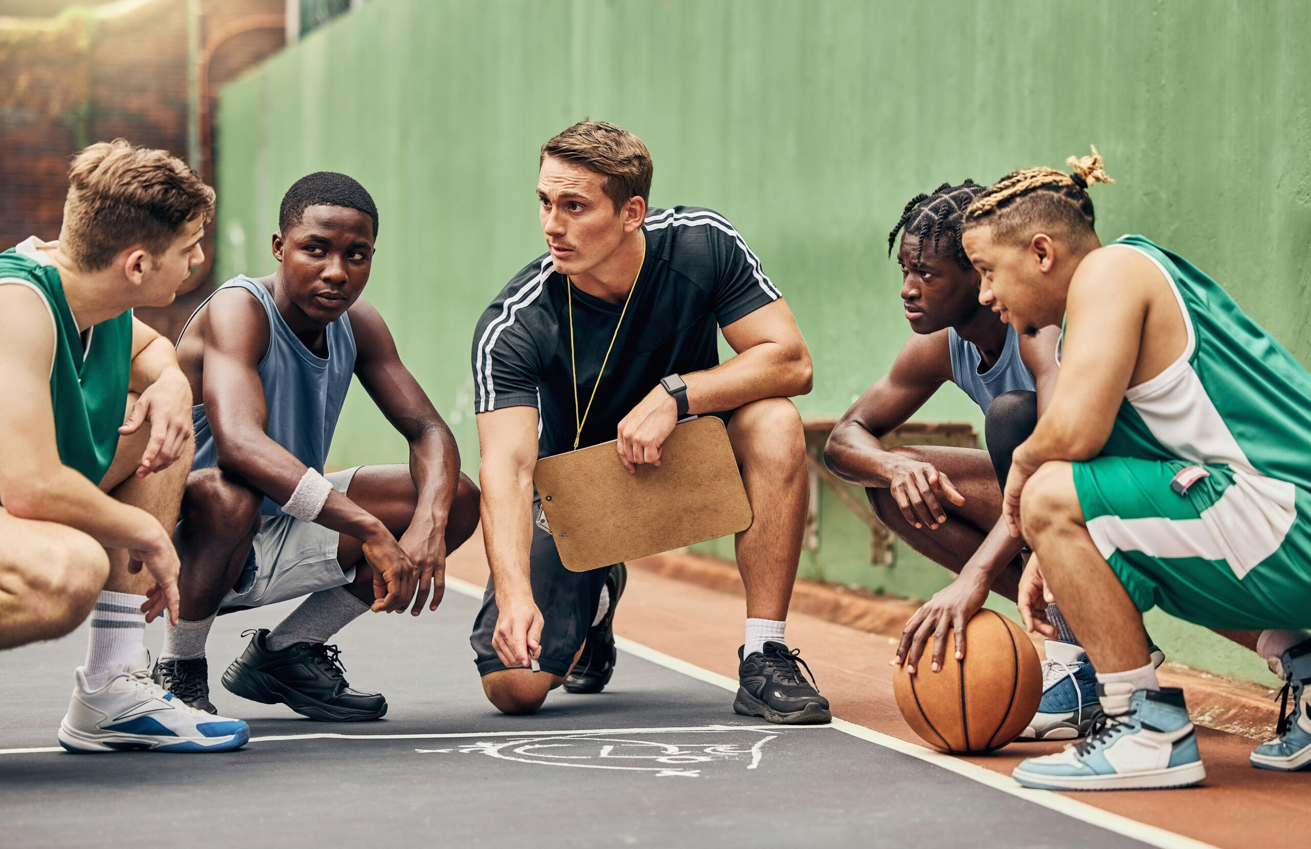 Ein Trainer erkl&auml;rt Taktiken an einem Basketballplatz, umgeben von f&uuml;nf Spielern in Sportkleidung.