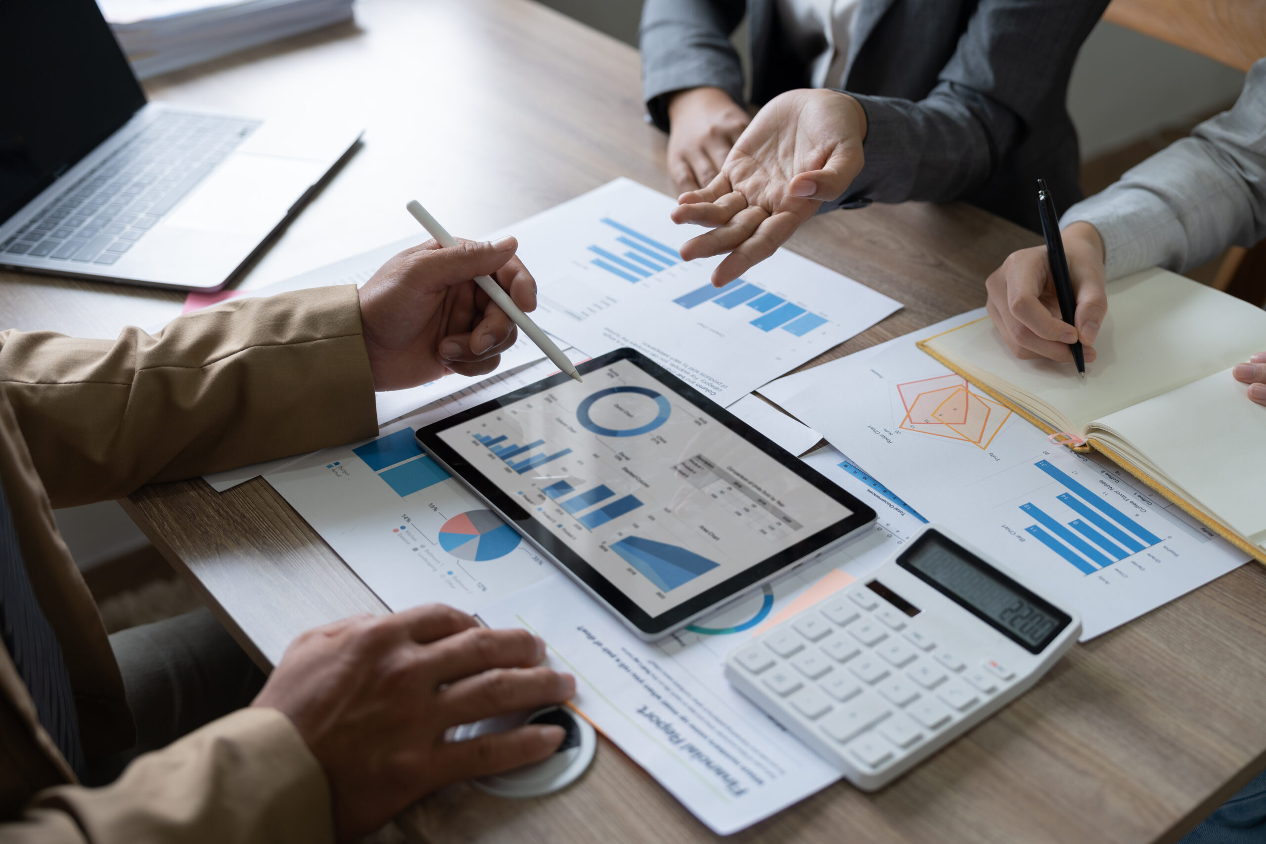 Besprechung mit Diagrammen und einem Tablet, w&auml;hrend Teilnehmer Daten analysieren und Ideen austauschen.